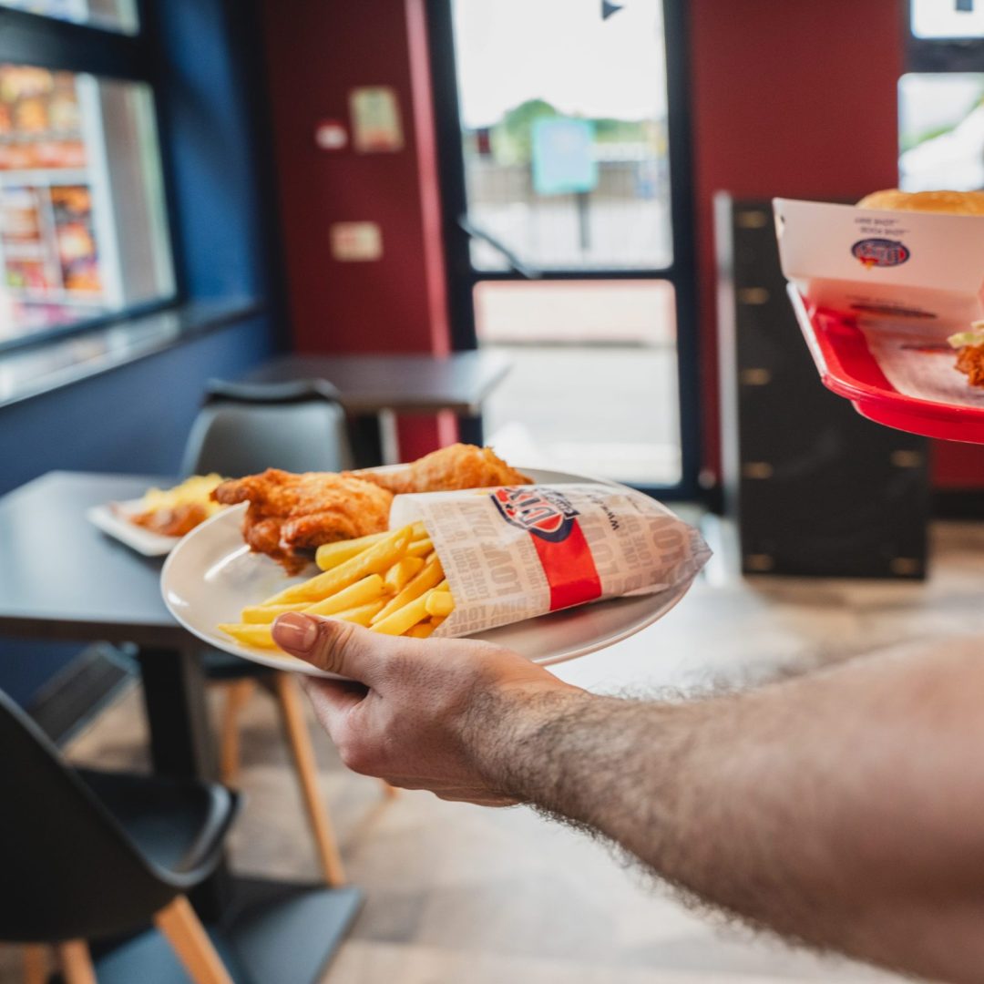 Hand carrying a tray with Dixy Chicken fried food and fries, with restaurant interior in the background.